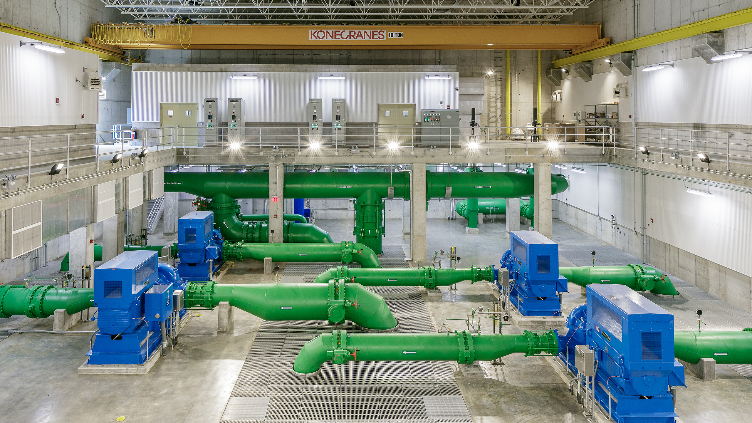 Large industrial room with green pipes and blue machinery on the floor, overhead crane labeled “Konecranes,” control panels along the back wall, and metal guardrails on a raised walkway.