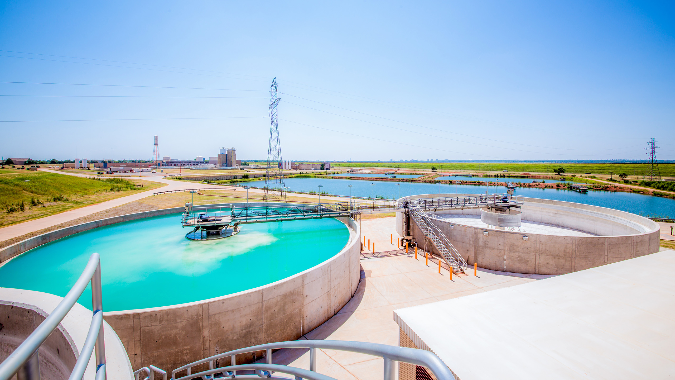 A modern water treatment facility with large circular tanks filled with blue water, surrounded by grassy fields, power lines, and several buildings under a clear blue sky.