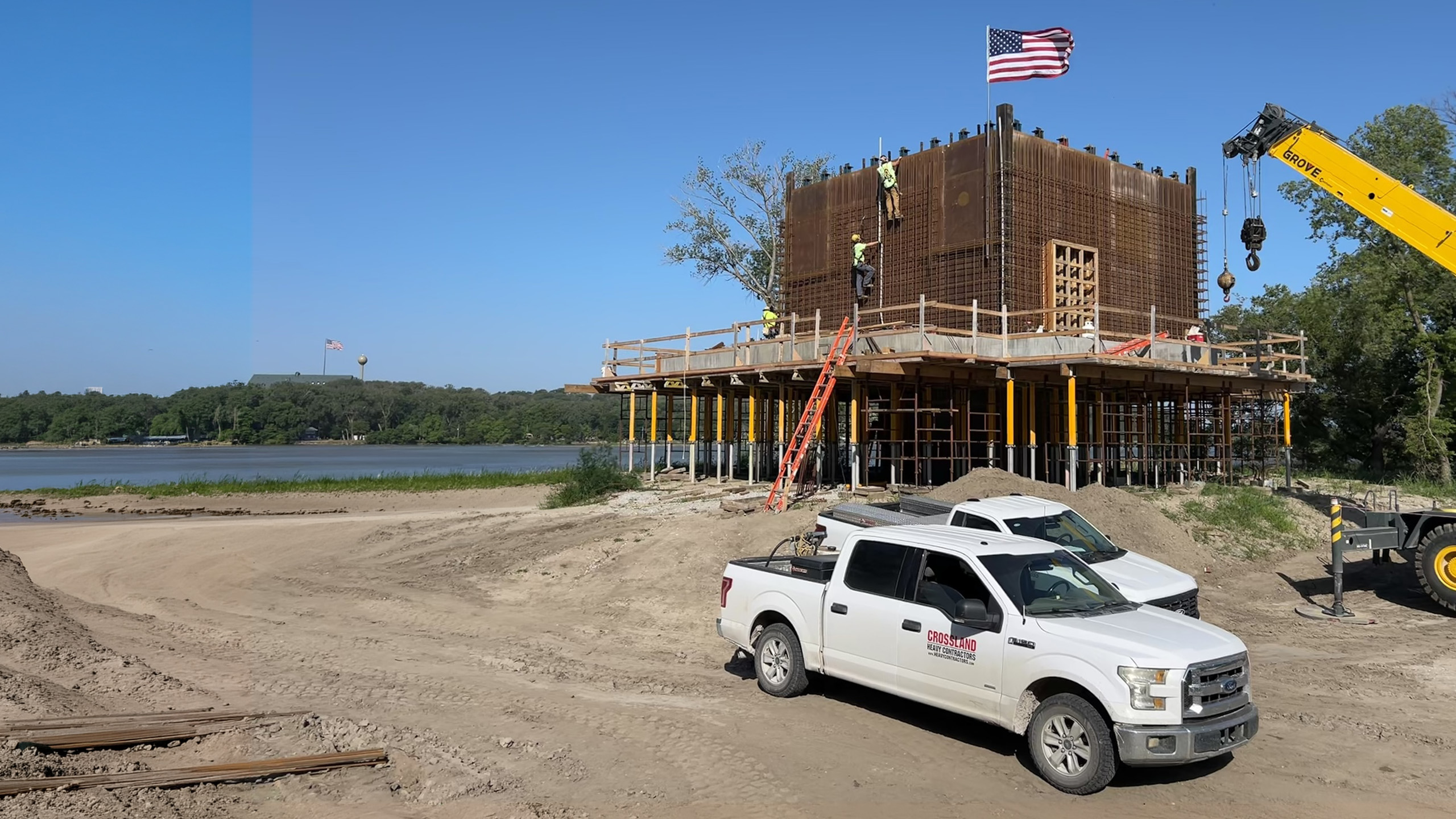 Construction site by a lake with workers on a large, boxy steel structure, a raised American flag, a crane, two white trucks, and sandy ground. Trees and water are visible in the background under a clear sky.