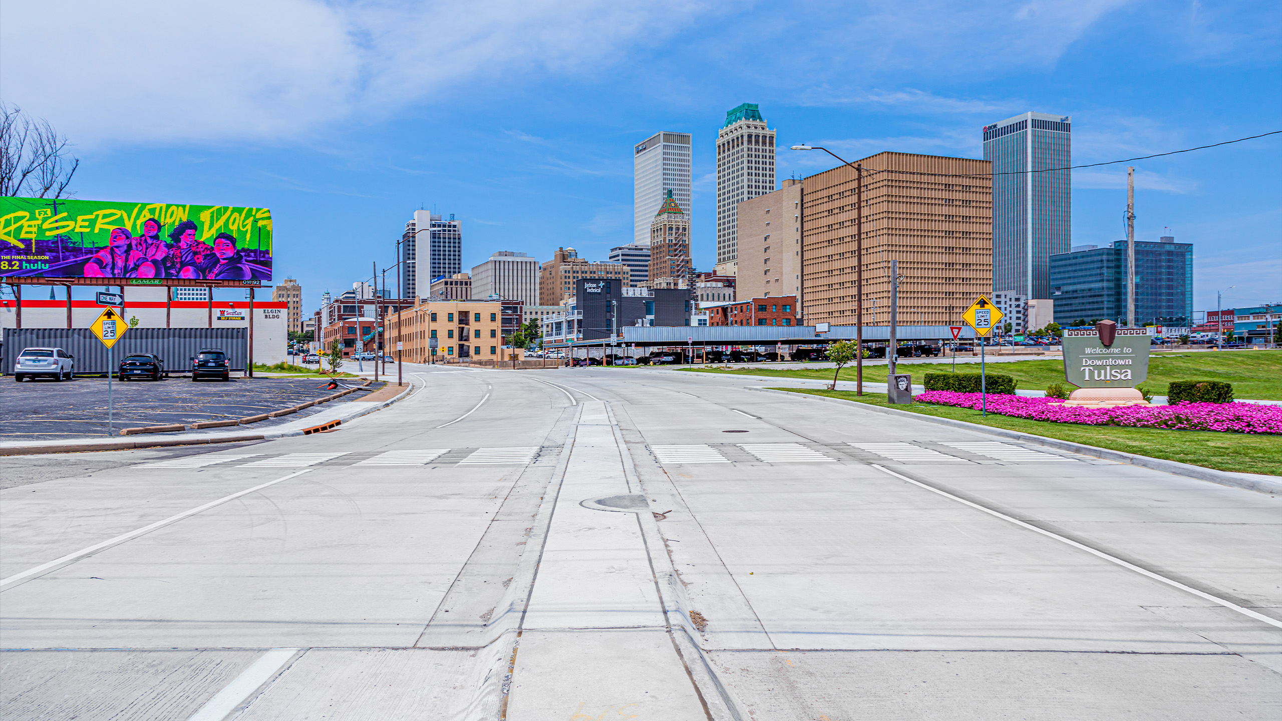 A wide, empty street leads into downtown Tulsa with tall buildings, a green “Reservation Dogs” billboard, bright pink flowers, and a “Tulsa” sign on the right under a partly cloudy sky.