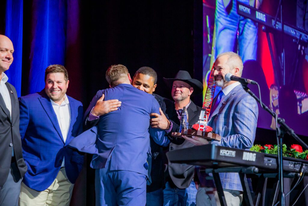 A group of men in suits, some smiling, stand on stage as one hugs another. One man holds a tray with a colorful trophy. Stage lights and a keyboard are visible, with a large projected screen in the background.