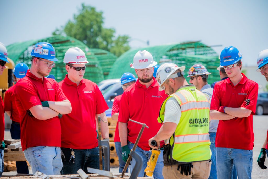 A group of construction workers in red shirts and hard hats watch a worker in a safety vest demonstrate equipment at a worksite outdoors.