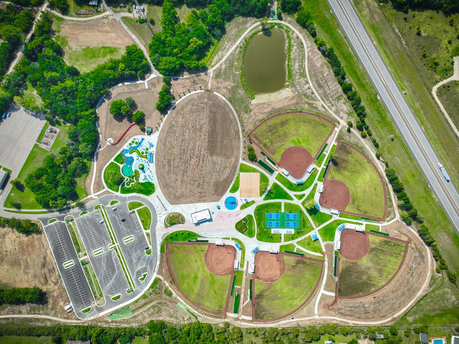 Aerial view of a large park featuring multiple baseball fields, tennis and basketball courts, a splash pad, a playground area, parking lots, walking paths, grassy areas, and a pond; bordered by a highway and trees.