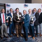 Eight men in suits and dress clothes stand in front of an "ABC Arkansas" backdrop, smiling and holding multiple eagle-shaped awards and a plaque, appearing at an awards event or ceremony.