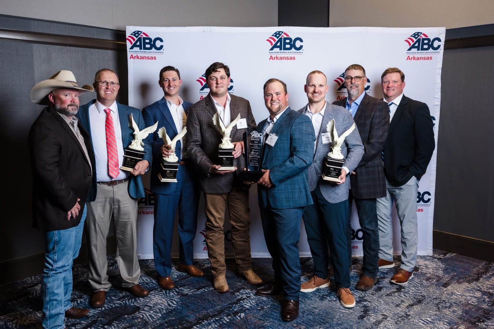 Eight men in suits and dress clothes stand in front of an "ABC Arkansas" backdrop, smiling and holding multiple eagle-shaped awards and a plaque, appearing at an awards event or ceremony.