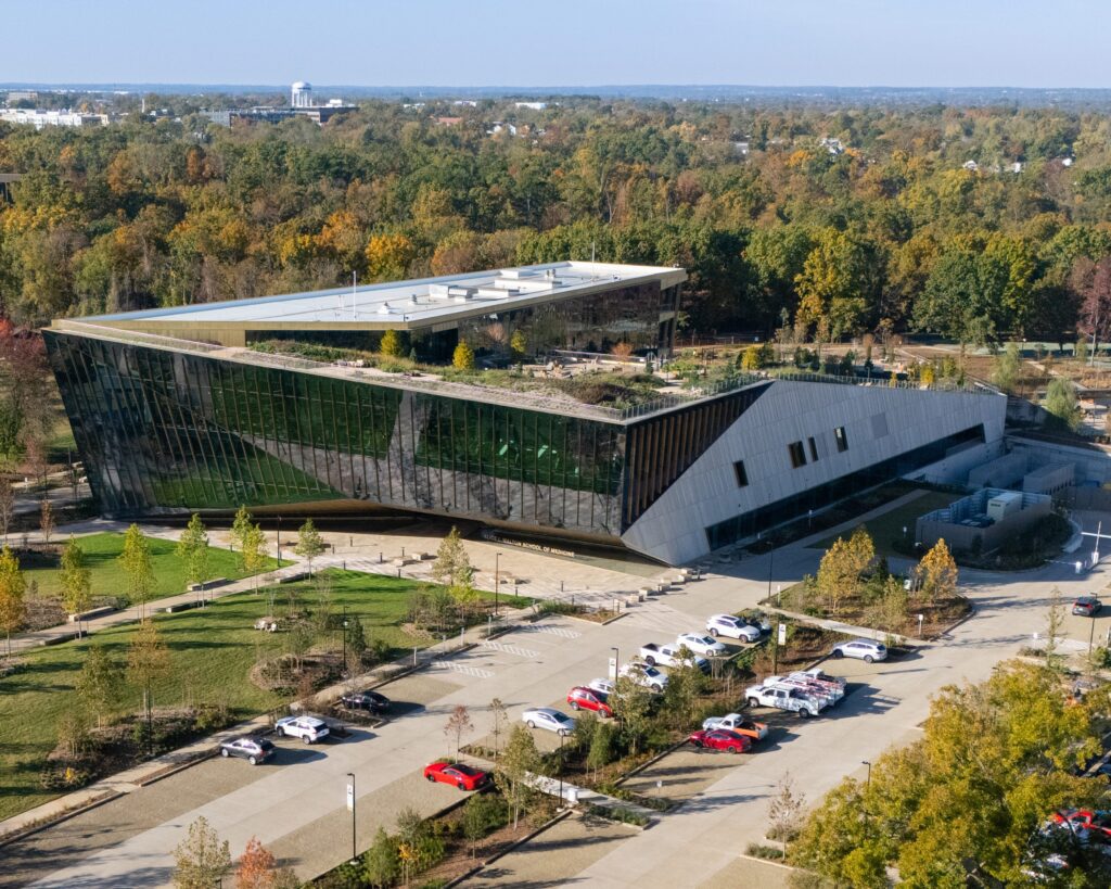 Aerial view of a modern building with angular design and reflective glass facade, surrounded by trees with fall foliage, landscaped grounds, and a parking lot with several cars.
