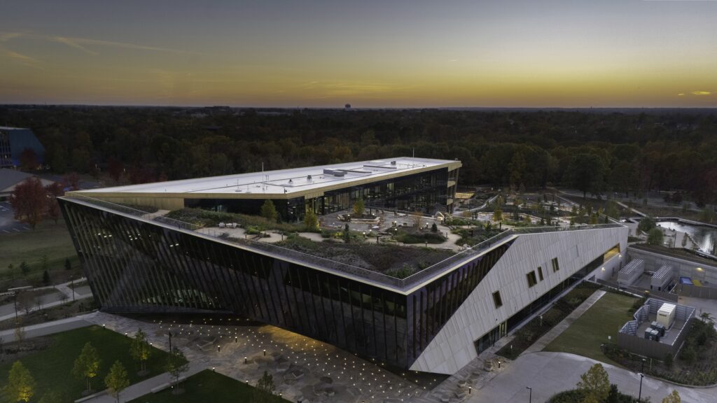 A modern, angular building with a green rooftop and large glass windows is shown at sunset, surrounded by trees and open green spaces. The sky is a gradient of orange and purple hues.