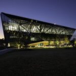 A modern, angular glass building with illuminated interiors stands surrounded by trees at dusk, with a darkening sky and a grassy area in the foreground.