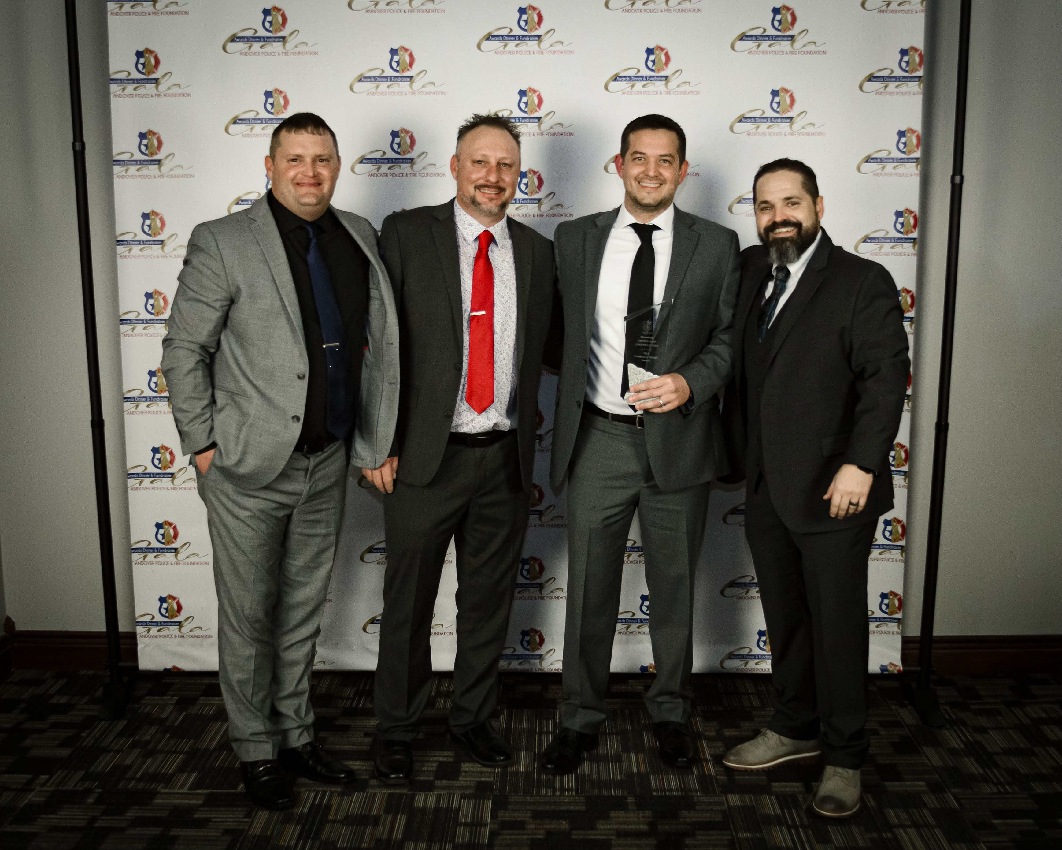 Four men in suits stand side by side, smiling at the camera. One man holds a glass award. They pose in front of a step-and-repeat backdrop with logos, on a patterned carpeted floor.
