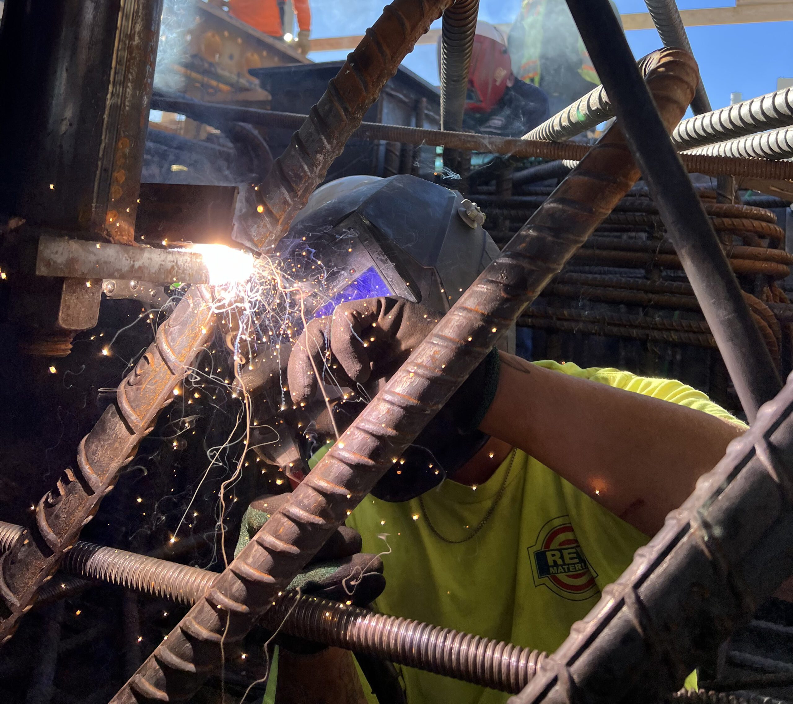 A welder wearing protective gear uses a welding torch on steel rebar, creating bright sparks. The worker is surrounded by metal rods and wears a yellow shirt with a visible logo.