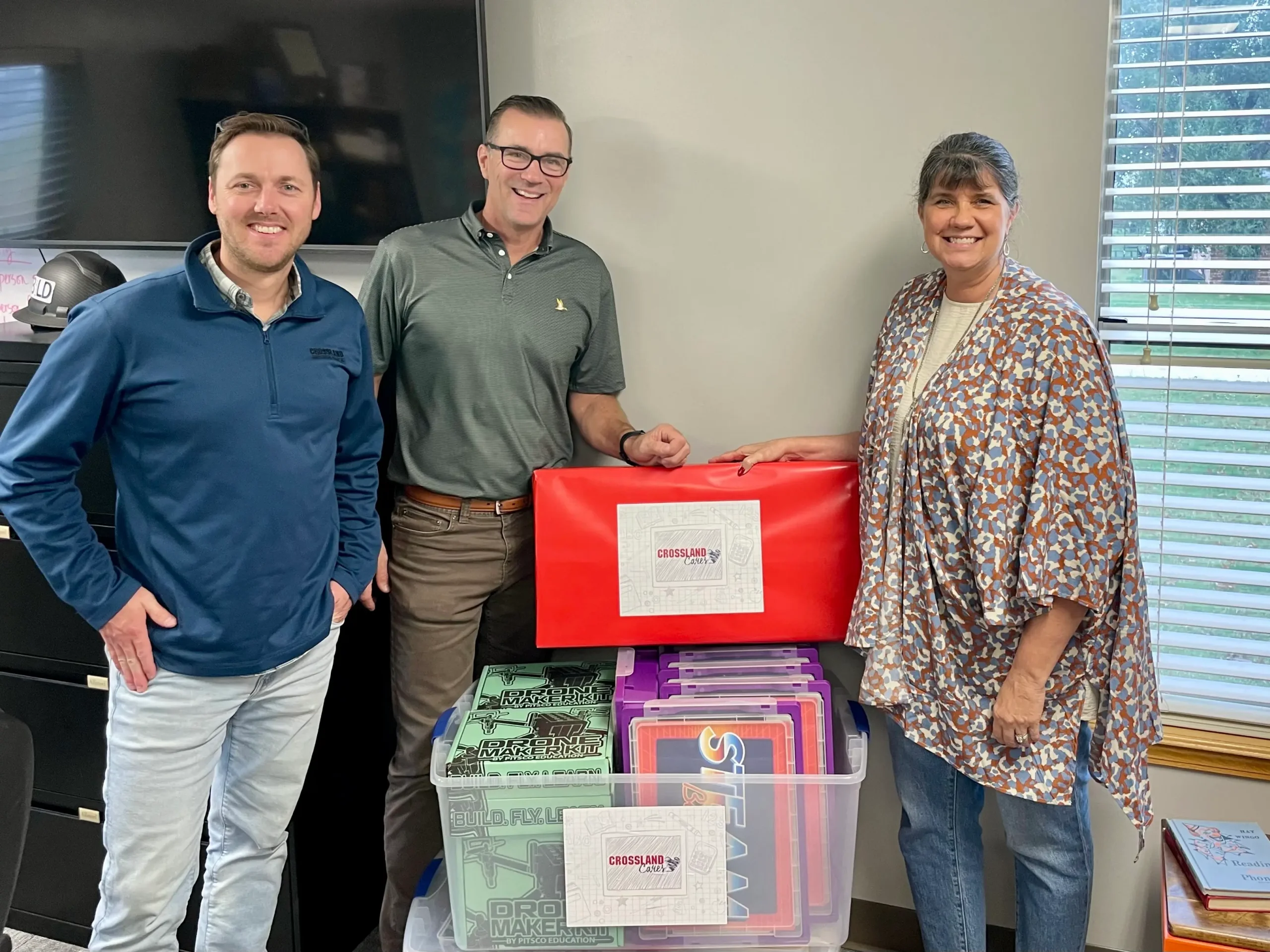 Three adults stand smiling in an office next to plastic bins and a large red package labeled “CROSSLAN LOVE.” Two men wear casual shirts and pants; a woman in a patterned shawl stands to the right.
