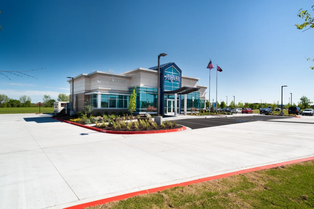 A modern car wash building with large windows sits in front of a landscaped area and parking lot. Two flagpoles display flags, and several cars are parked nearby under a clear blue sky.