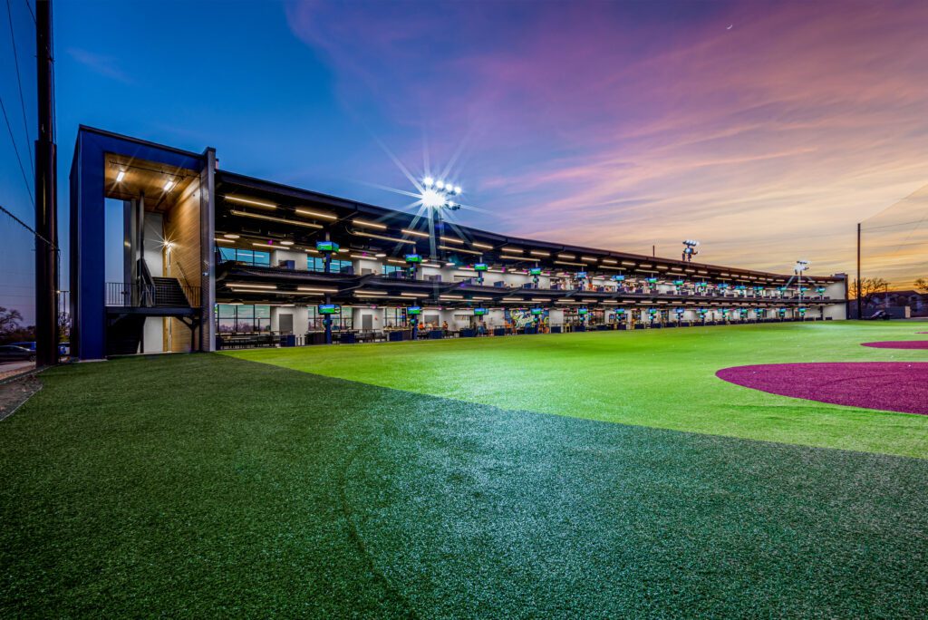 A modern multi-level driving range with bright lights overlooks a well-maintained artificial turf field at sunset, with a colorful sky in the background.