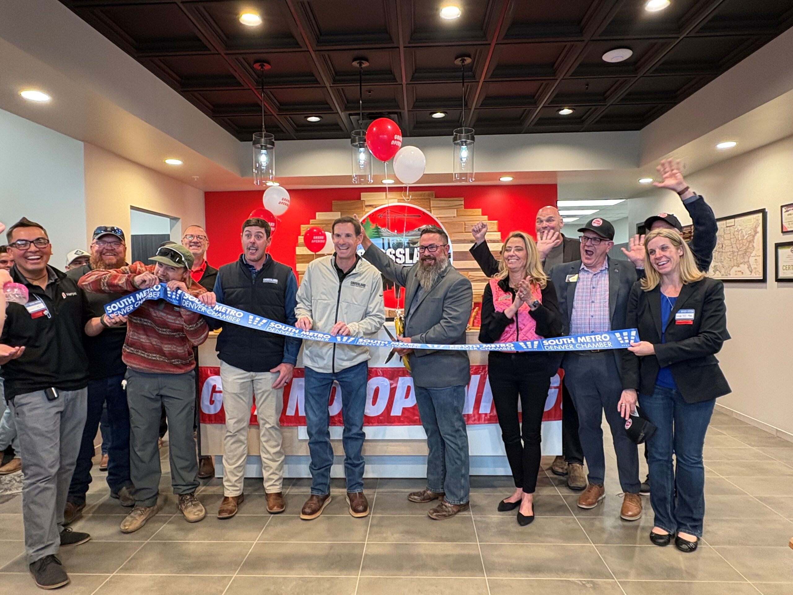 A group of people smile and cheer as they cut a blue ribbon at an indoor ribbon-cutting ceremony, with balloons and a red wall in the background.