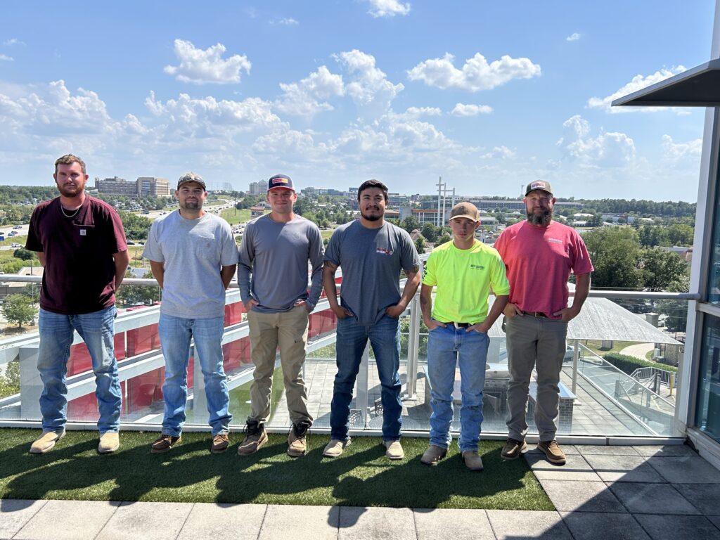 Six men stand in a row on a rooftop with a cityscape and blue sky in the background. They are dressed casually in jeans, t-shirts, and work boots, with some wearing caps. The mood appears relaxed and informal.