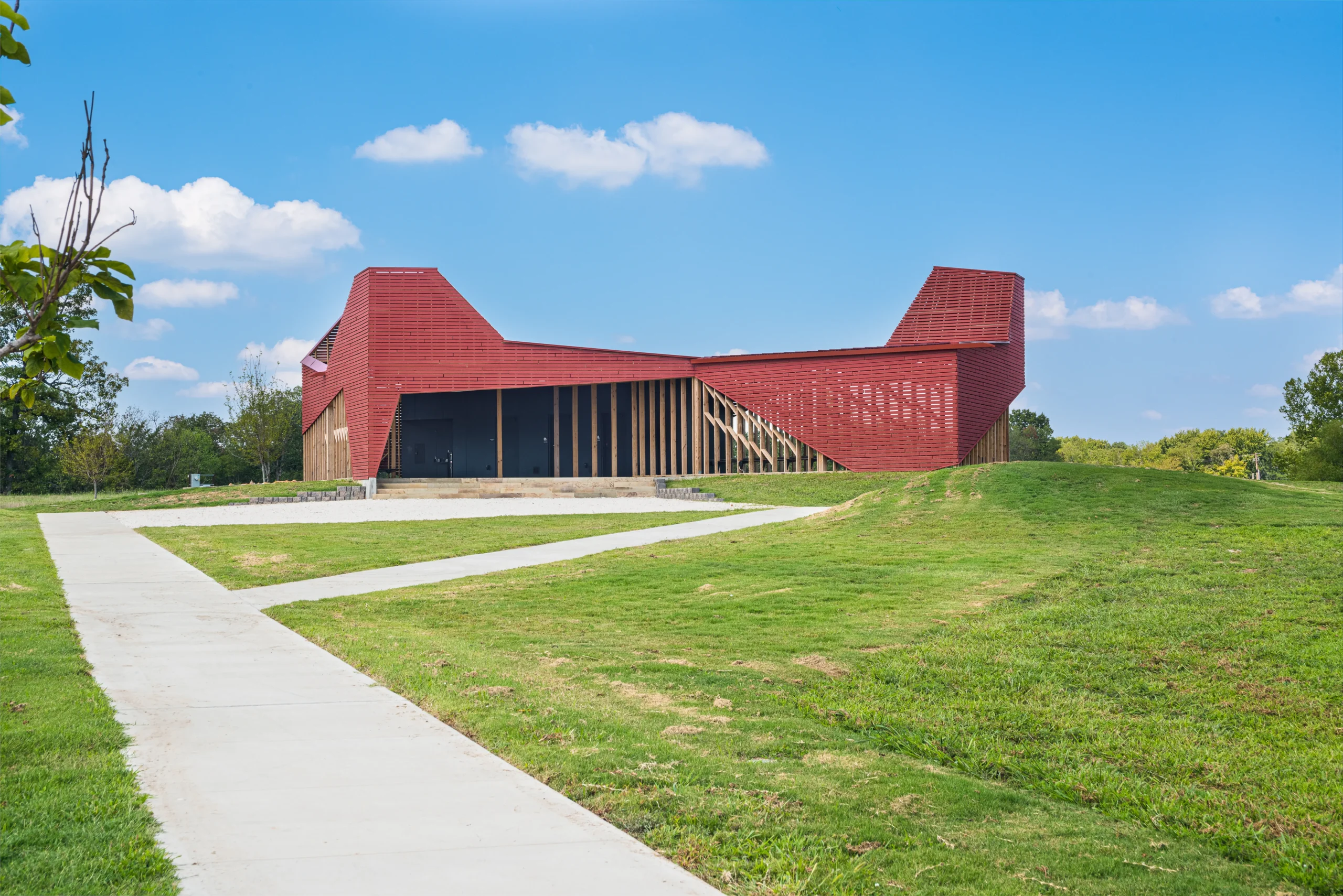 A modern red pavilion with angular architecture sits on a grassy hill under a blue sky with scattered clouds, with a concrete path leading up to the structure.