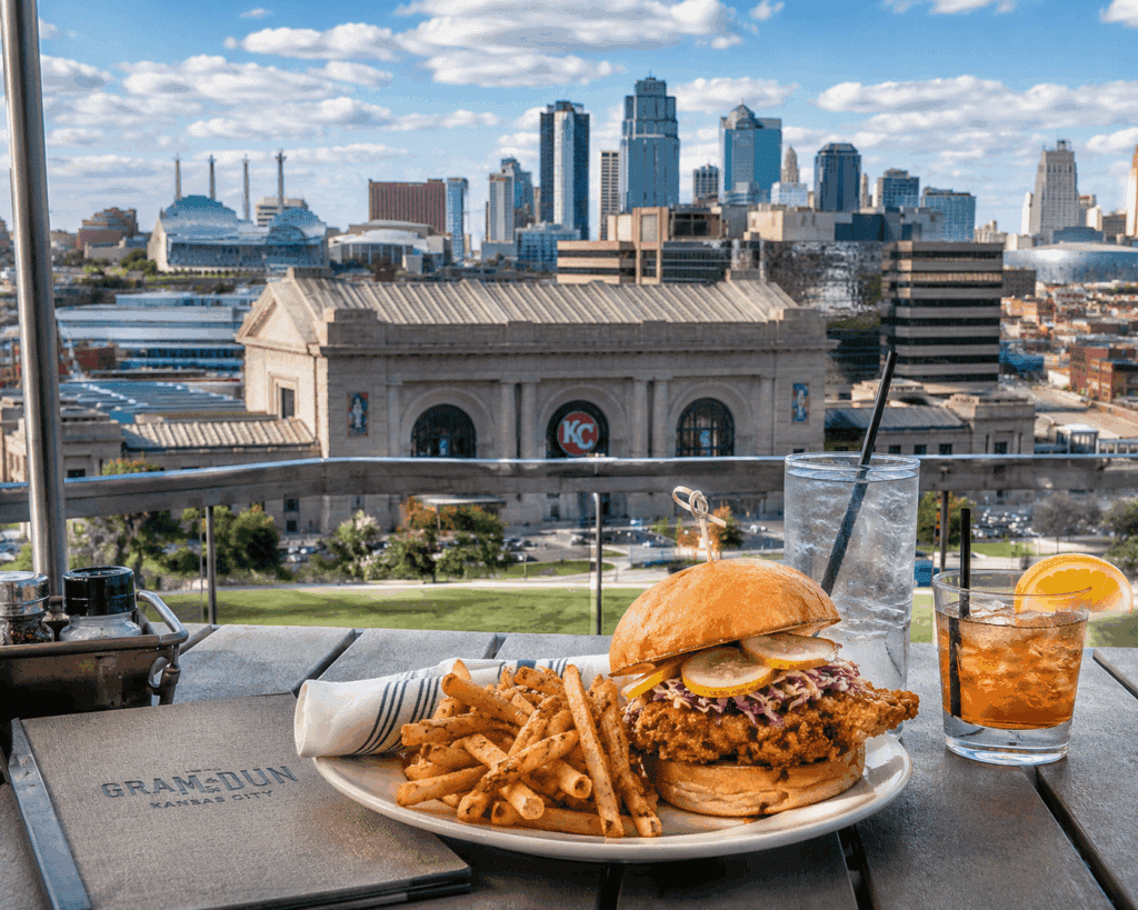 A crispy chicken sandwich with fries, iced tea, and water sits on a table overlooking the city skyline and the iconic "KC" sign. A menu labeled "GranDun" hints at what’s special about Crossland dining in this historic setting.