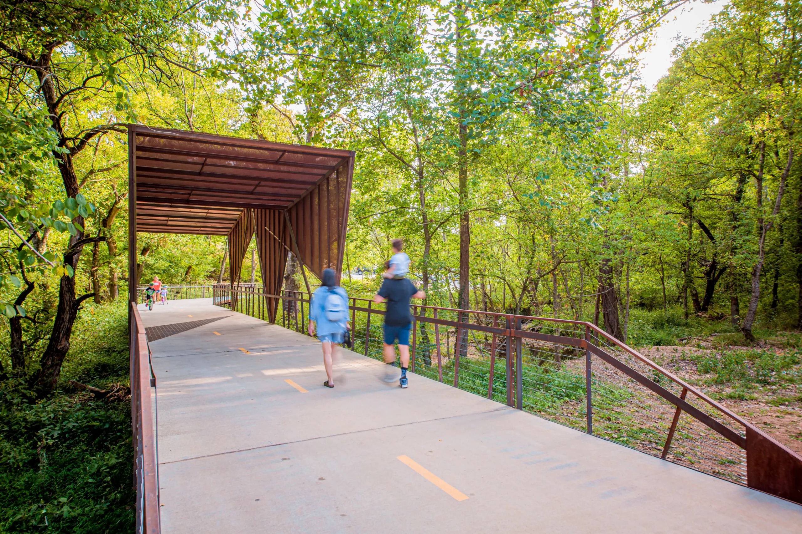 Two people walk on a paved trail through a green forest, approaching a modern, rust-colored metal bridge structure. More people are visible in the distance, enjoying the tree-lined path.