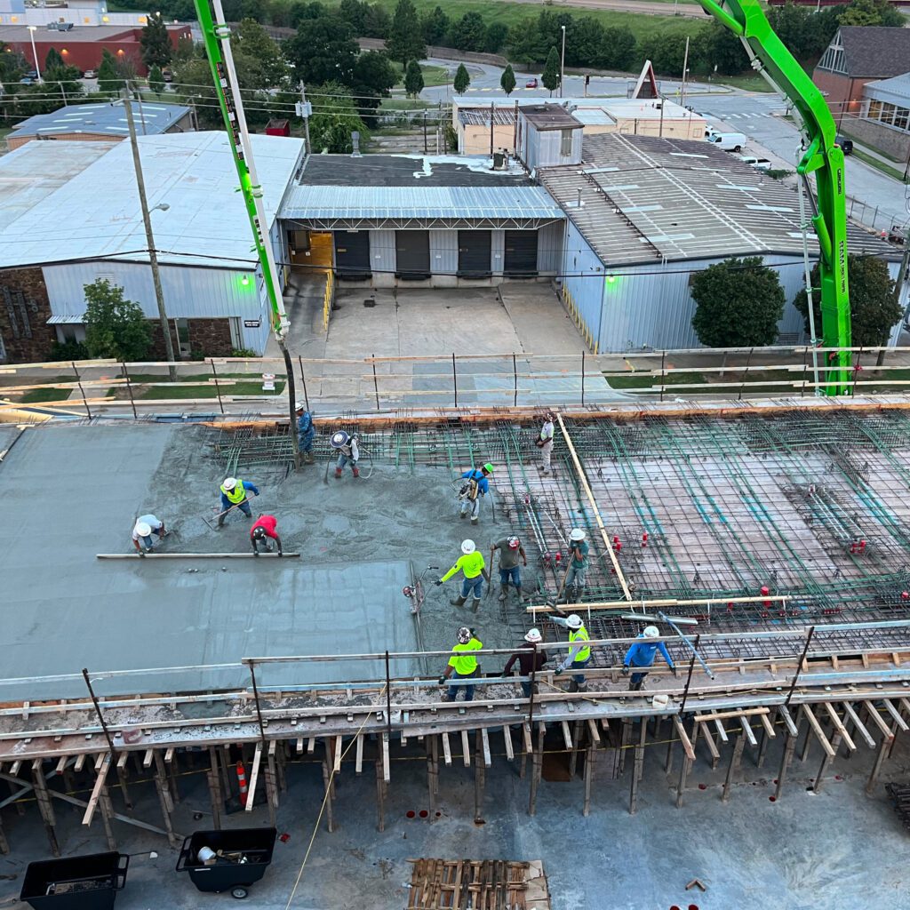 Construction workers wearing safety gear are smoothing freshly poured concrete on a building site. Some use tools while others supervise. Equipment and scaffolding surround the site, and industrial buildings are visible in the background.