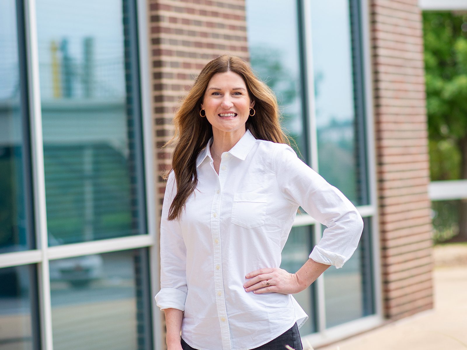 A woman with long brown hair, wearing a white button-up shirt, stands smiling with one hand on her hip in front of a building with large windows and brick walls.