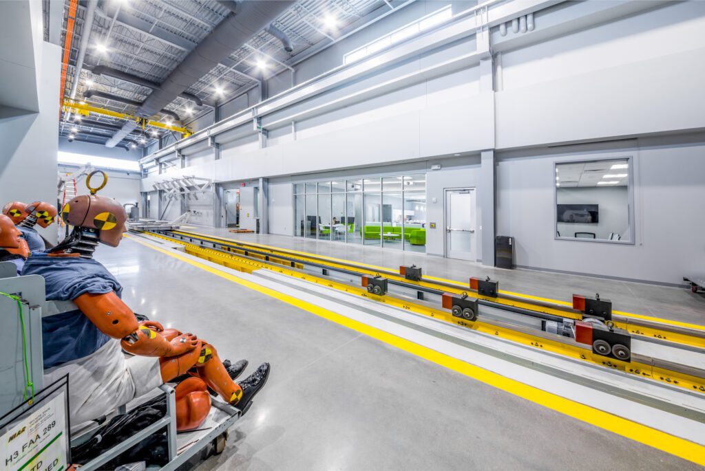 A brightly lit industrial facility with crash test dummies seated on a cart near long yellow testing tracks, adjacent to glass-walled offices and workspaces.