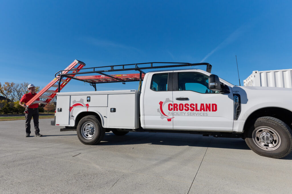 A white Crossland Facility Services truck is parked on a concrete surface. A worker in a red shirt and helmet stands nearby, holding a ladder from the truck’s roof rack under a clear blue sky.
