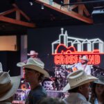 People in cowboy hats gather indoors at a country music venue. A musician performs on stage with instruments, and a neon “Crossland” sign with a guitar and silos glows in the background.
