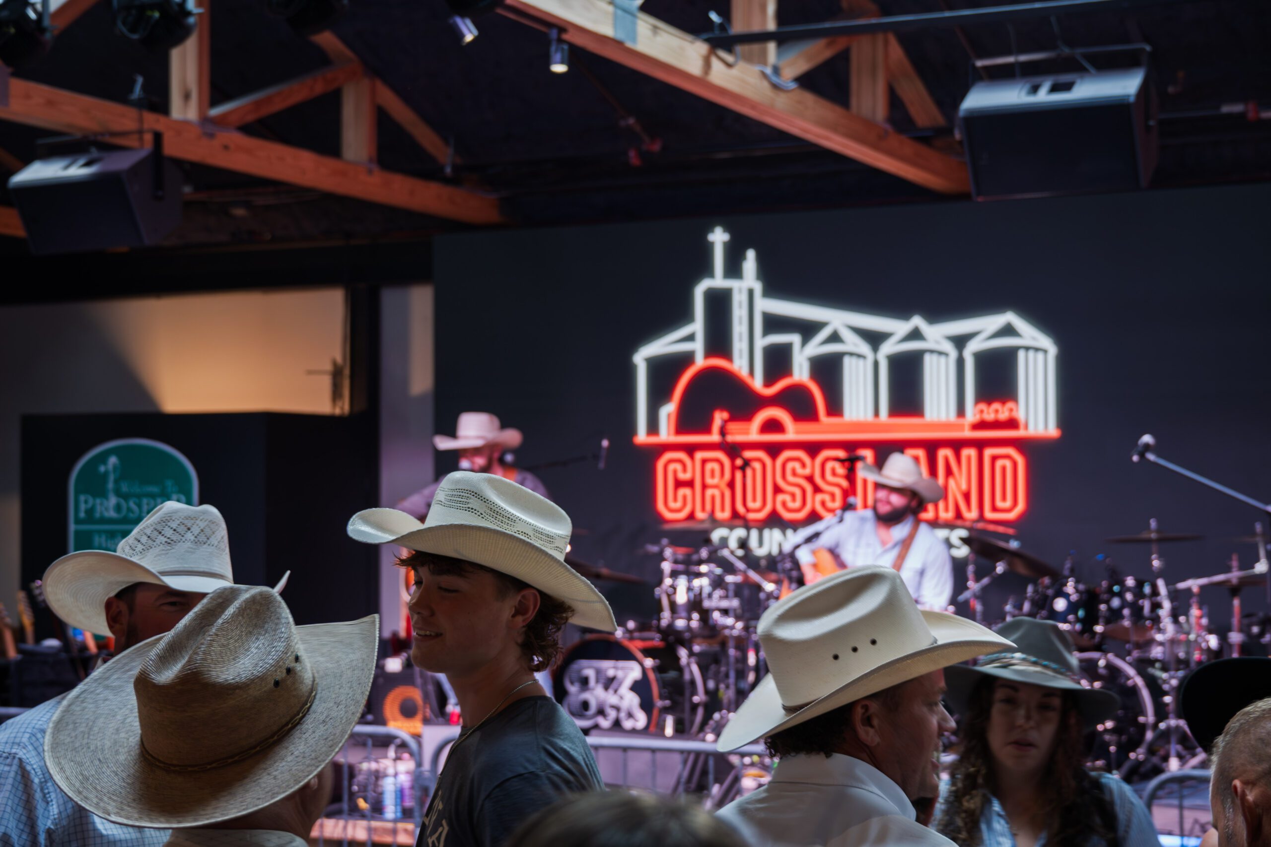 People in cowboy hats gather indoors at a country music venue. A musician performs on stage with instruments, and a neon “Crossland” sign with a guitar and silos glows in the background.