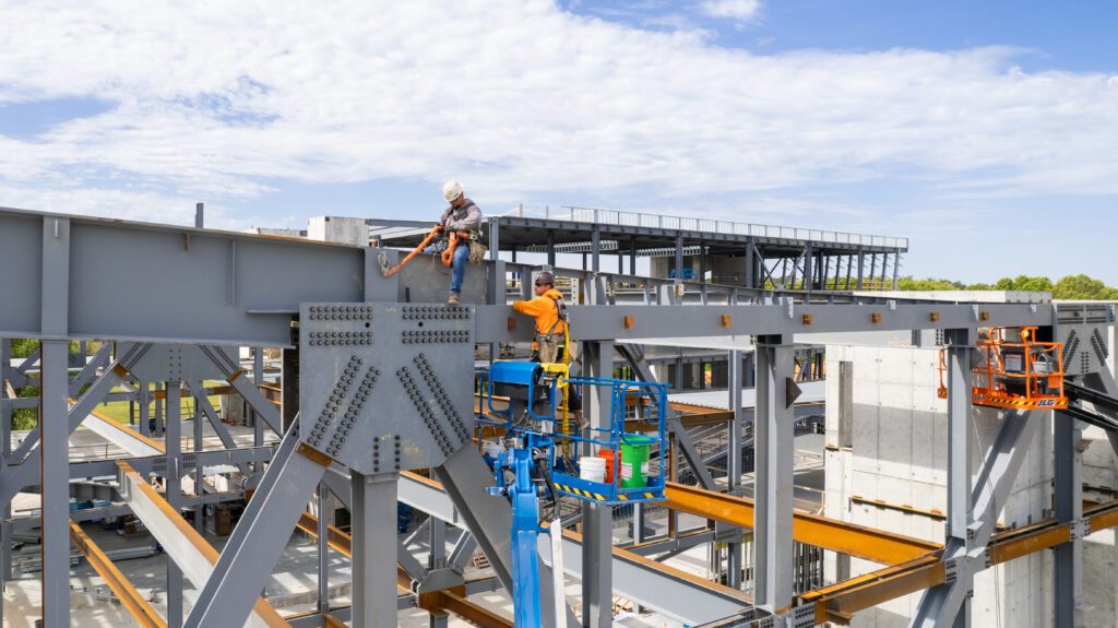 Two construction workers wearing safety gear work on assembling a steel framework, with one standing on the structure and the other operating a boom lift under a partly cloudy sky.