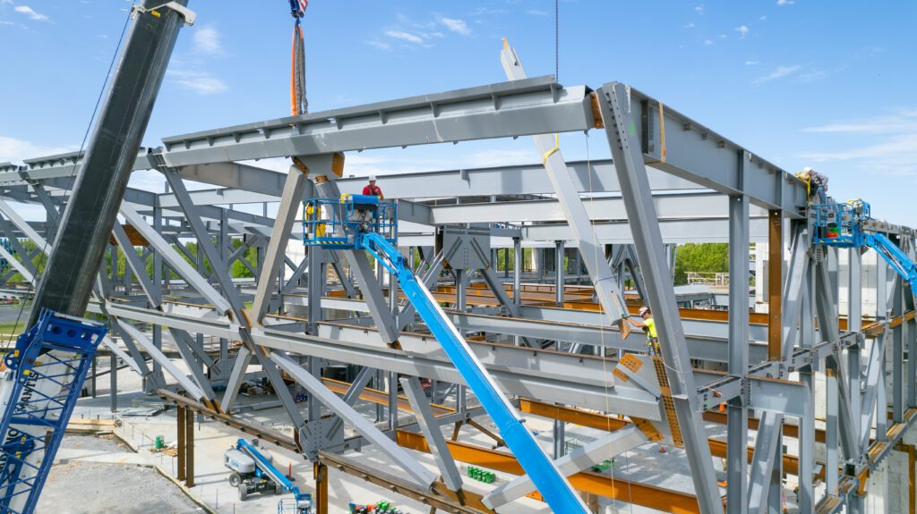 Construction workers on lifts are assembling a large steel framework for a building under a clear blue sky, with cranes and beams visible around the structure.