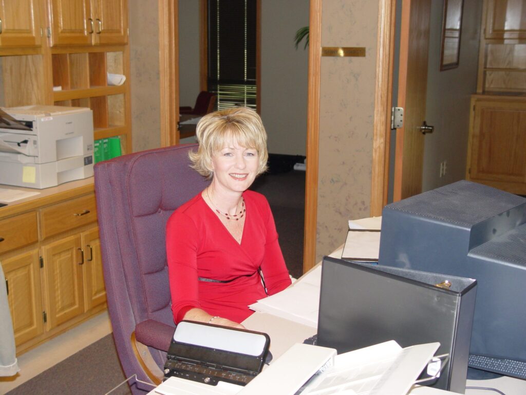 A woman with short blond hair, wearing a red dress, sits and smiles at a desk in an office with wooden cabinets, papers, and a computer monitor.