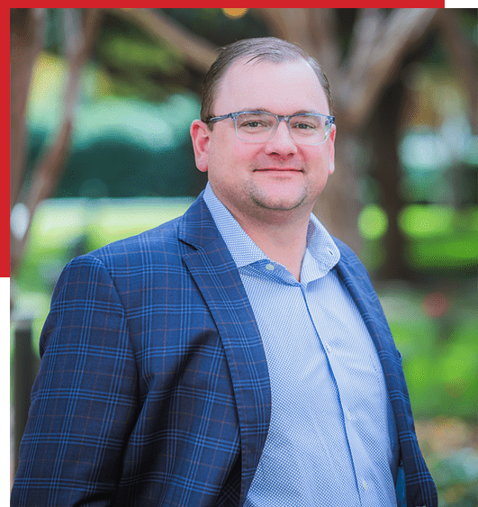 A man in glasses and a blue plaid suit jacket stands outdoors, smiling slightly. The background is blurred greenery with trees, and the image has a partial red border on the top and left sides.
