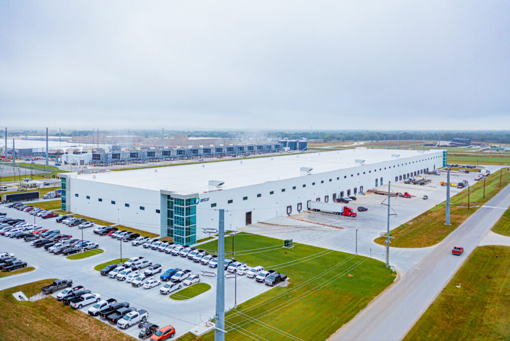A large industrial warehouse with a white roof sits beside a parking lot filled with cars and trucks on a cloudy day, with roads and grassy areas surrounding the building.