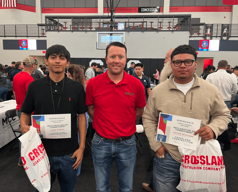 Three men stand indoors at an event. The man in the center, wearing a red polo, is flanked by two young men holding certificates and gift bags. People and tables are visible in the background.