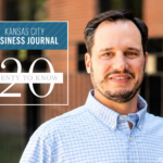 A man in a light blue checkered shirt stands outside. Overlay text reads: “Kansas City Business Journal 20 Twenty to Know.” Brick building and black fence are in the blurred background.