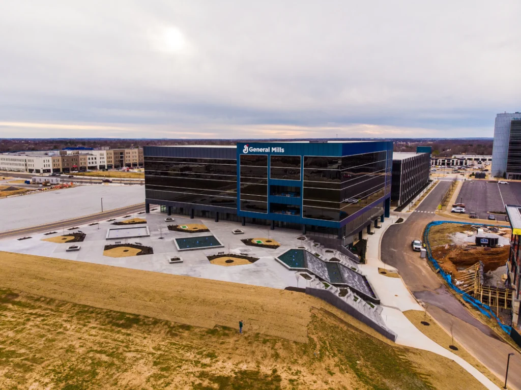 Aerial view of a modern, glass-fronted General Mills office building with landscaped areas and parking lots, surrounded by roads, construction, and nearby buildings under a cloudy sky.