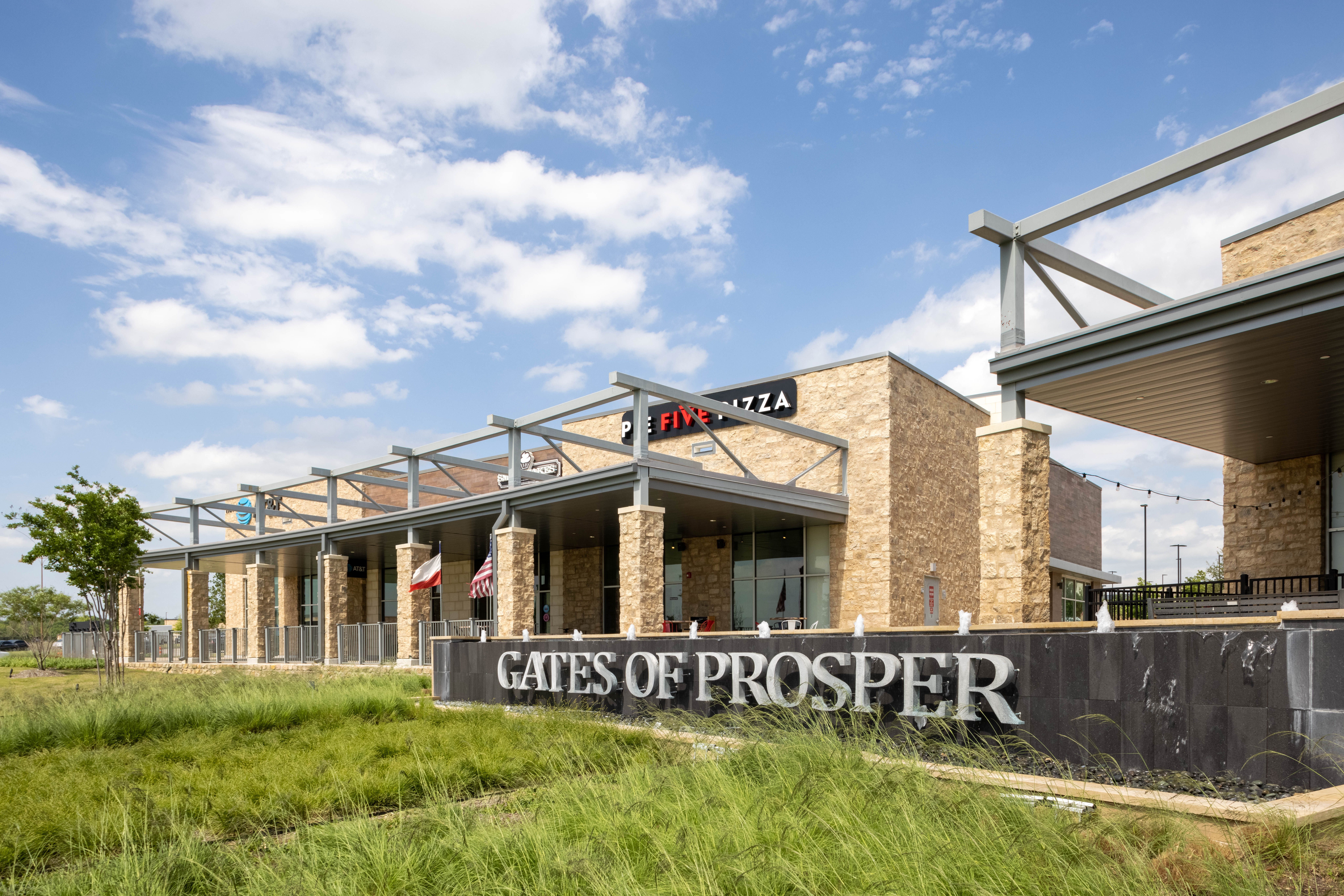A modern shopping center with stone buildings and a sign reading "Gates of Prosper" in front, set against a blue sky with clouds and landscaped grass in the foreground.