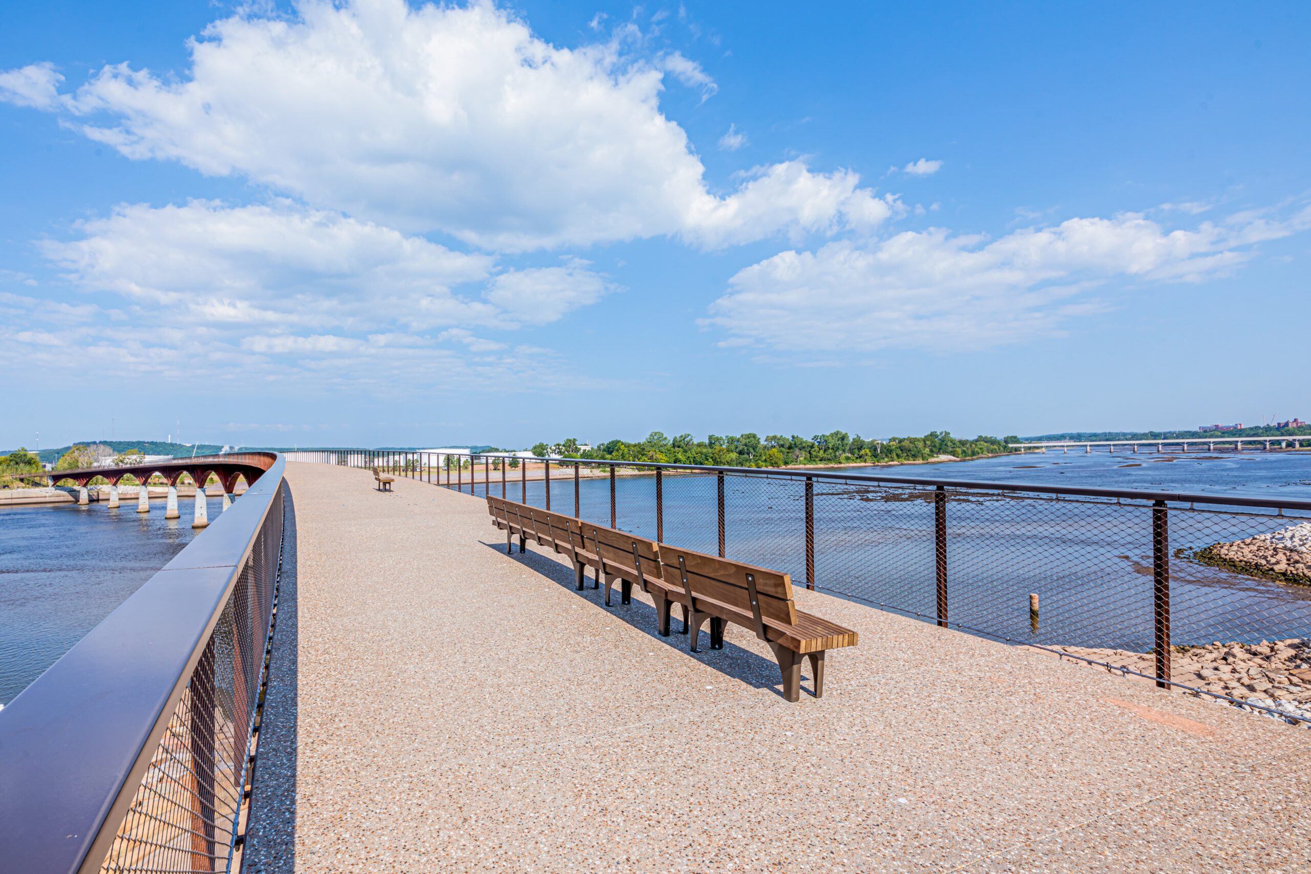 A wide pedestrian bridge with benches crosses over a river on a sunny day, with blue sky, scattered clouds, and trees in the background.