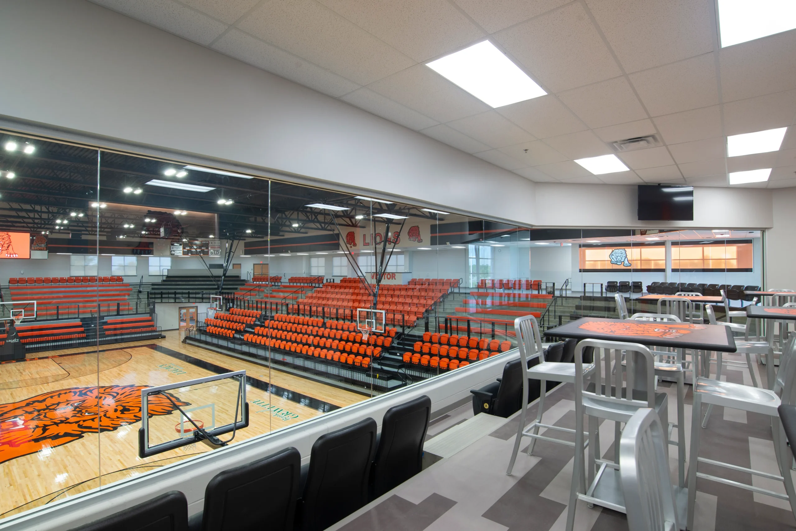 A modern indoor basketball court with orange and black seating, viewed from a glass-walled lounge area with high tables and chairs. The gym is well-lit with a large tiger graphic at center court.