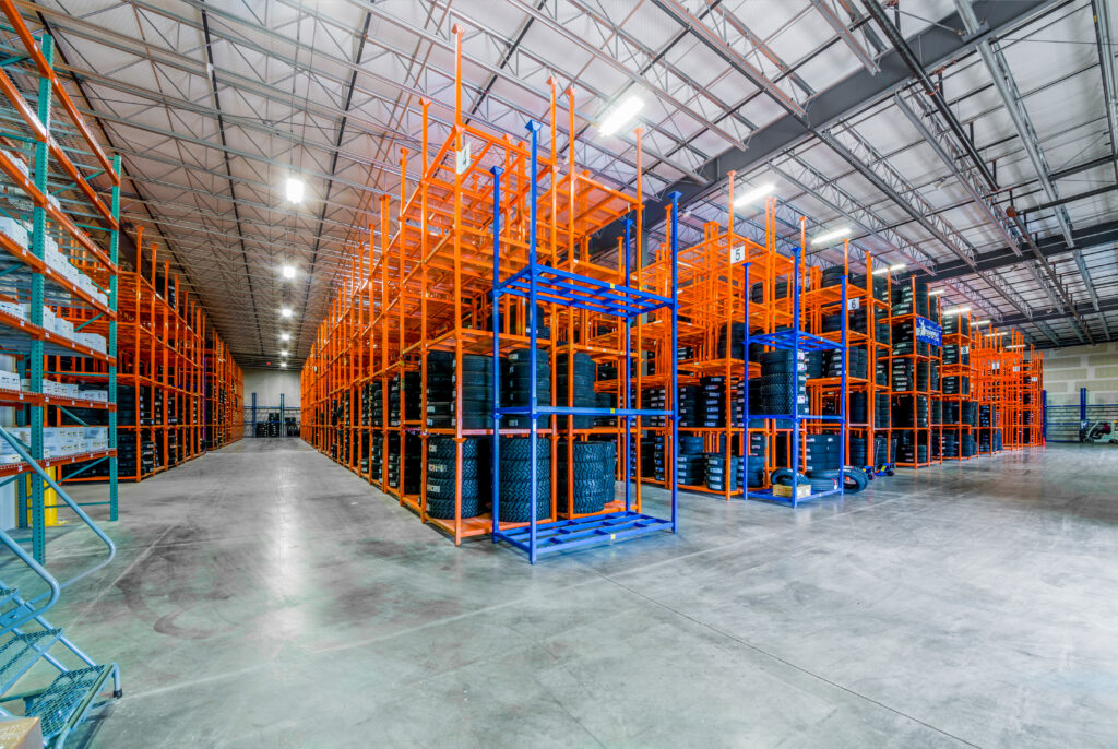 Large warehouse interior with tall, empty orange and blue metal shelving units, concrete floor, and bright overhead lighting. The space is organized and spacious, with wide aisles and a high ceiling.
