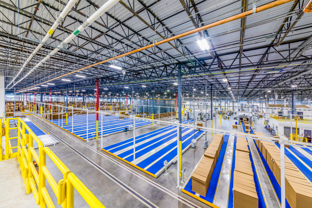 Large warehouse interior with tall, empty orange and blue metal shelving units, concrete floor, and bright overhead lighting. The space is organized and spacious, with wide aisles and a high ceiling.