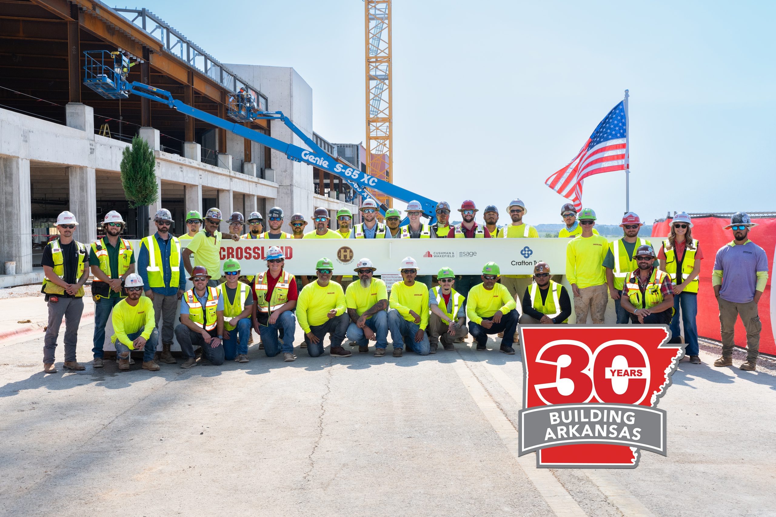 A large group of construction workers in safety gear pose in front of a building site, holding a beam. An American flag waves in the background. A “30 Years Building Arkansas” badge is in the lower right corner.