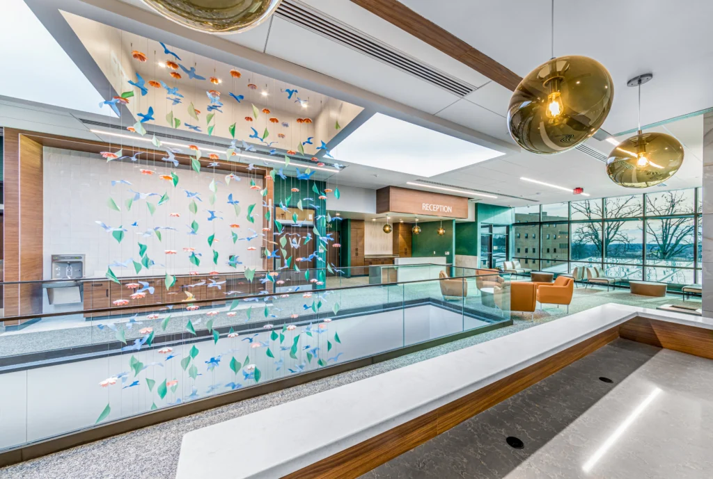 Modern reception area with a suspended colorful bird sculpture, marble counters, tan chairs, and large windows showing trees outside; round golden pendant lights hang from the ceiling.
