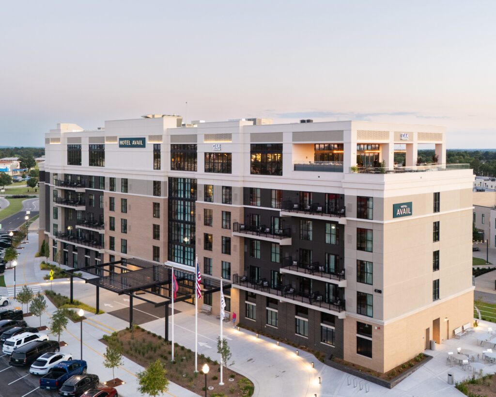 A modern, multi-story hotel building with balconies, large windows, and Hotel Avail signs on the upper floors, surrounded by parked cars, landscaping, and flagpoles at the entrance.