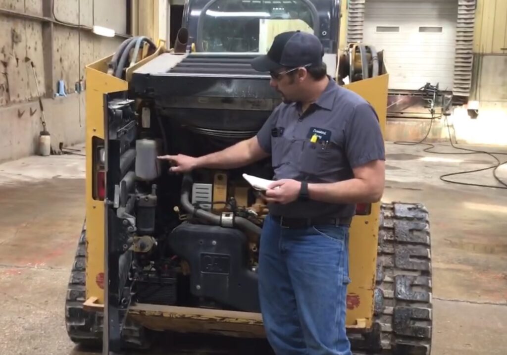 A man in work clothes points at machinery components inside the open rear compartment of a yellow tracked skid-steer loader in a workshop. He is holding a notepad and inspecting the equipment.