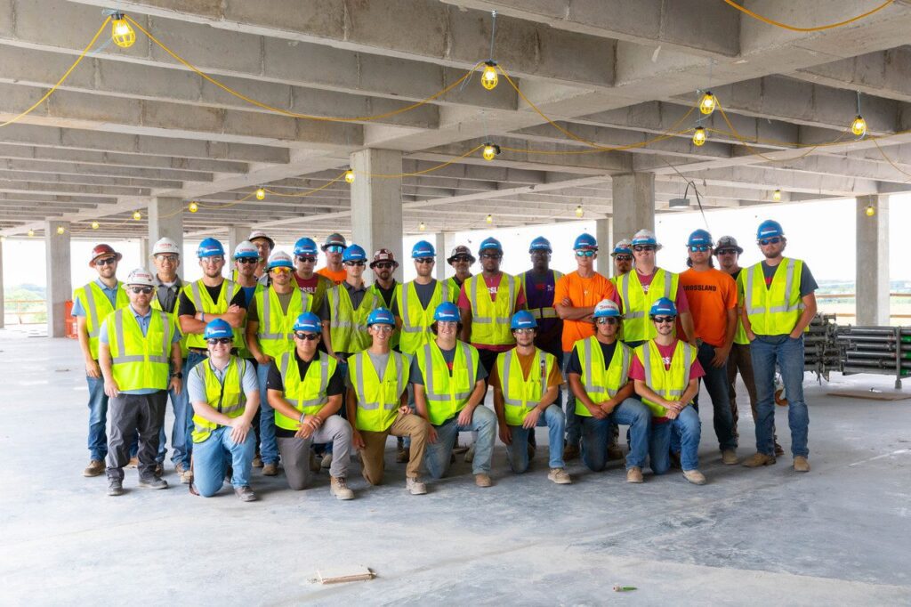 A group of construction workers wearing safety vests and hard hats pose for a photo inside an unfinished building with exposed ceilings and hanging lights.