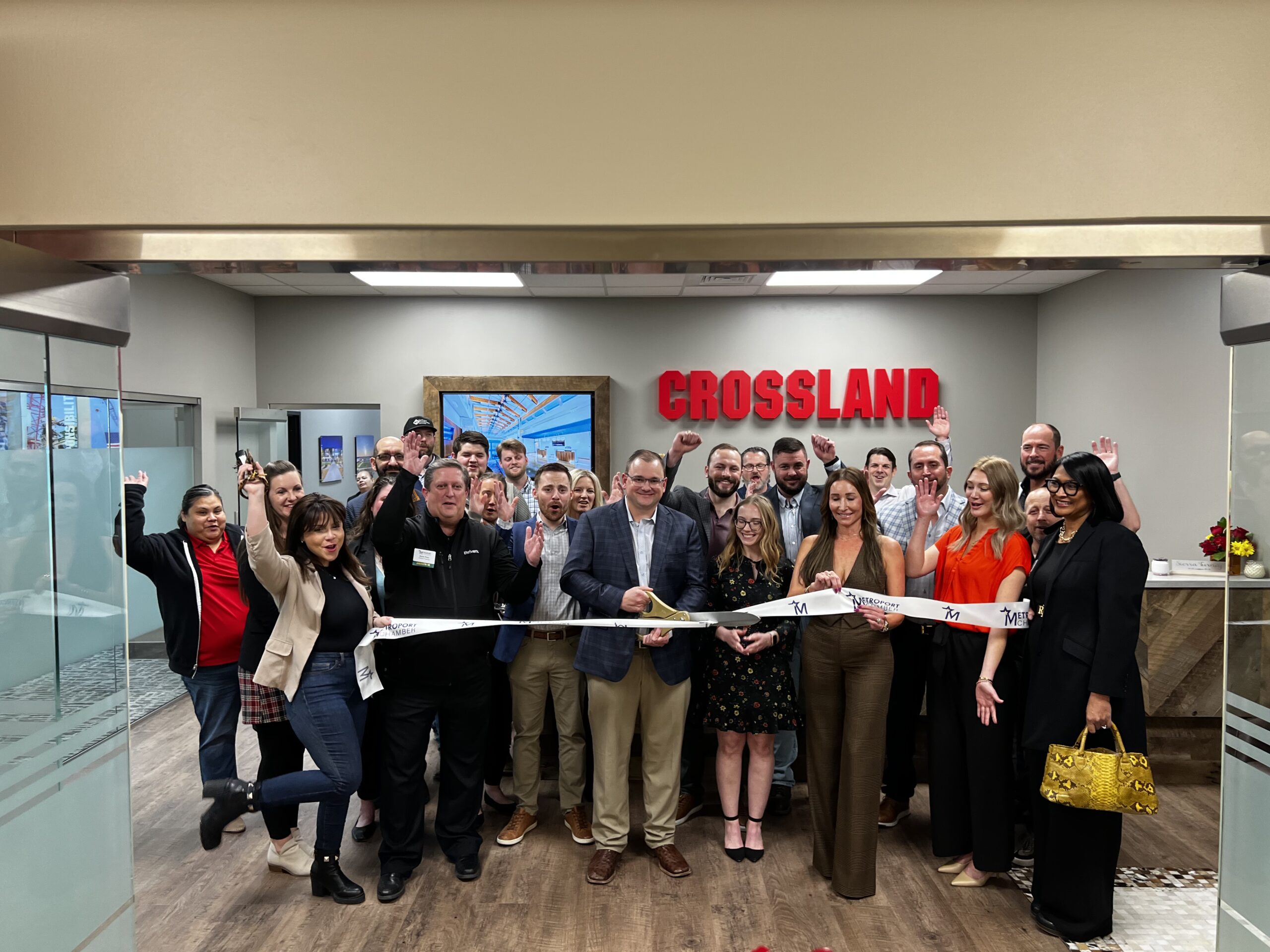 A large group of people stand inside an office, smiling and holding a ribbon for a ribbon-cutting ceremony. A sign reading "CROSSLAND" is on the wall behind them. Several people wave at the camera.