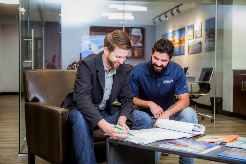Two men sit in an office, smiling and reviewing blueprints on a coffee table covered with magazines. One man is writing with a pencil as the other leans in to look. Glass walls and office furniture are in the background.