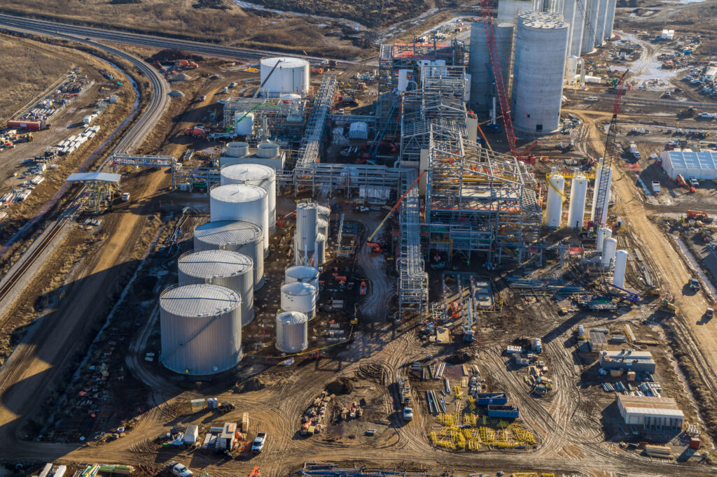 Aerial view of an industrial facility with large cylindrical storage tanks, tall silos, construction cranes, and scattered machinery at a dirt-covered site.
