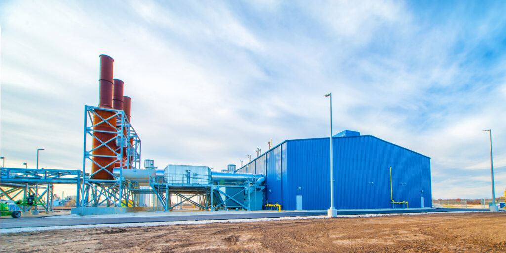 A large blue industrial building with three tall red chimneys, pipes, and equipment sits on a dirt and concrete lot under a cloudy sky.